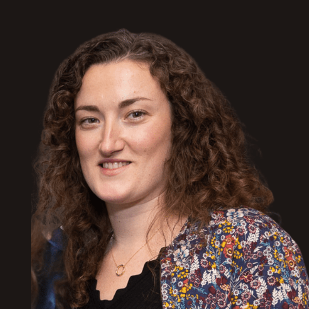 A woman with curly brown hair smiles slightly. She is wearing a floral-patterned jacket over a black top and a gold necklace, evoking the vibrant spirit of Ealaín na Gaeltachta. The background is solid black.