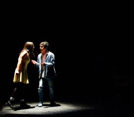 Two people stand under a spotlight on a dark stage, engaged in an intense conversation. Highlighting Ealaín na Gaeltachta, the woman wears a yellow dress and boots, whilst the man is dressed in casual jeans and a shirt.