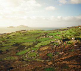 A scenic view of rolling green hills, part of the Ealaín na Gaeltachta landscape, dotted with small houses and fields, leading to the coastline where two small islands rest under a partly cloudy sky.