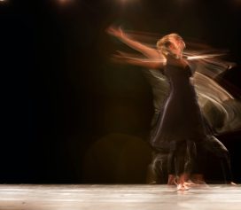 A dancer in a black dress performs on stage with blurred motion, creating a dynamic effect that evokes the expressive artistry of Ealaín na Gaeltachta against a dark background under soft stage lighting.
