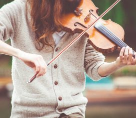 A person with long hair, wearing a beige cardigan, plays a violin outdoors. The focus is on their hands and the violin, evoking the spirit of Ealaín na Gaeltachta against a beautifully blurred background.