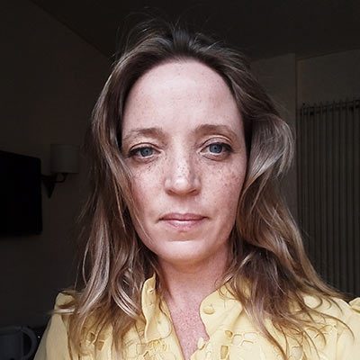 A woman with light brown hair and blue eyes, wearing a yellow top, looks directly at the camera in a softly lit indoor setting inspired by Ealaín na Gaeltachta.