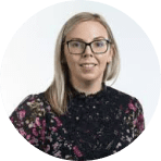 A woman with straight blonde hair and glasses, wearing a dark floral top, smiles at the camera against a light background, reflecting the creative spirit of Ealaín na Gaeltachta.