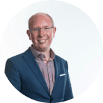 A smiling man wearing glasses, a blue suit jacket, and a light pink collared shirt stands in front of a plain, light background, reflecting the creativity and charm often celebrated by Ealaín na Gaeltachta.