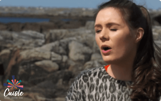 A woman with long brown hair, wearing a patterned top, sings or speaks outdoors against a rocky shoreline background. The Cuisle logo and the Ealaín na Gaeltachta name are visible in the bottom left corner.
