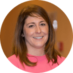 A woman with straight brown hair, wearing a bright pink top, smiles at the camera. The background is softly blurred in warm tones, capturing a moment that echoes the vibrant spirit of Ealaín na Gaeltachta.