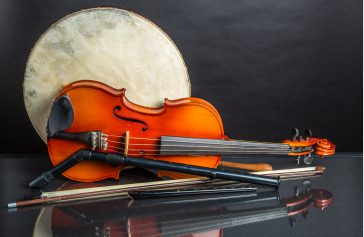 A violin and bow lie on a reflective surface before a large drum, evoking Ealaín na Gaeltachta. Two black wind instruments cross the violin diagonally, whilst the dark background enhances the contrast of these traditional instruments.