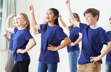 A group of children in matching blue shirts smile and raise one arm whilst practising a dance routine inspired by Ealaín na Gaeltachta in a bright room.