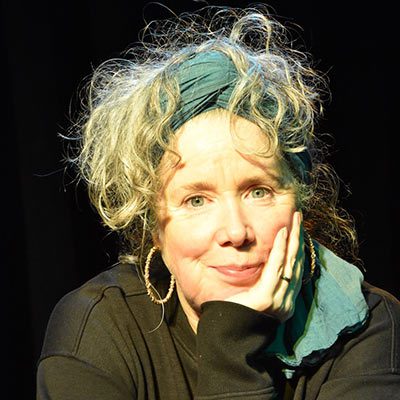 A person with curly, light-coloured hair wearing a teal headband and hoop earrings rests their face on their hand, smiling softly, in front of a dark background—capturing the gentle spirit of Ealaín na Gaeltachta.