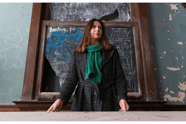 A woman with long brown hair, wearing a green scarf and grey coat, stands in front of a worn chalkboard covered with diagrams and the words beehive process—an inspiring scene reflecting the spirit of Ealaín na Gaeltachta in a weathered room.