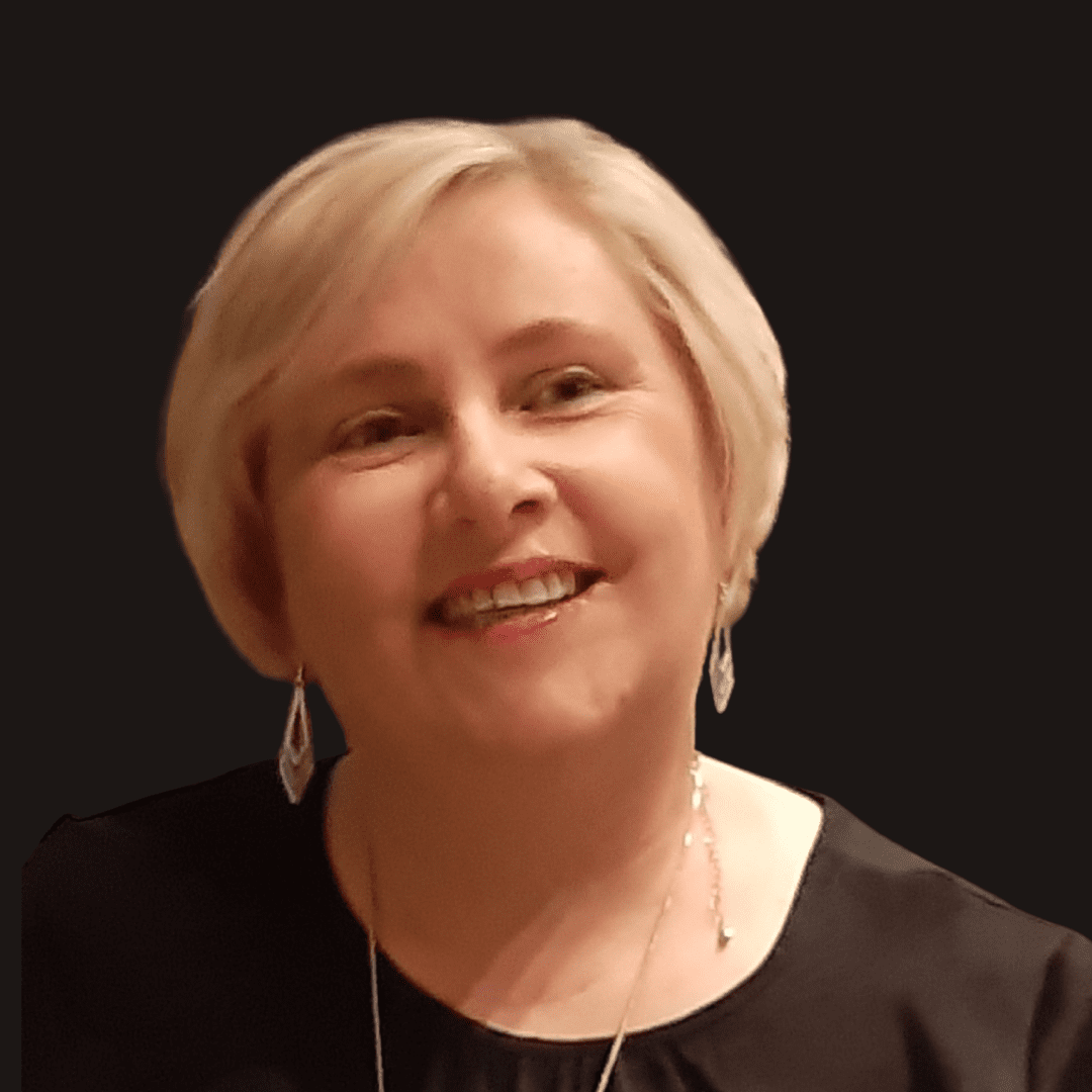 A smiling woman with short blonde hair wears dangling earrings and a black top, set against a plain dark background—her style subtly reflecting the creative spirit of Ealaín na Gaeltachta.