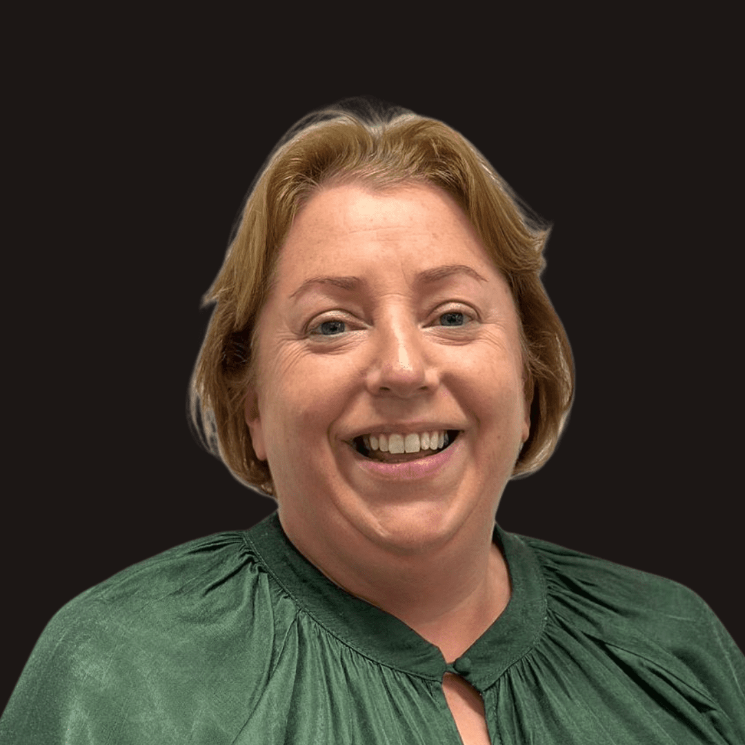 A smiling woman with light brown hair wearing a green blouse stands in front of a plain black background, reflecting the creative spirit of Ealaín na Gaeltachta.