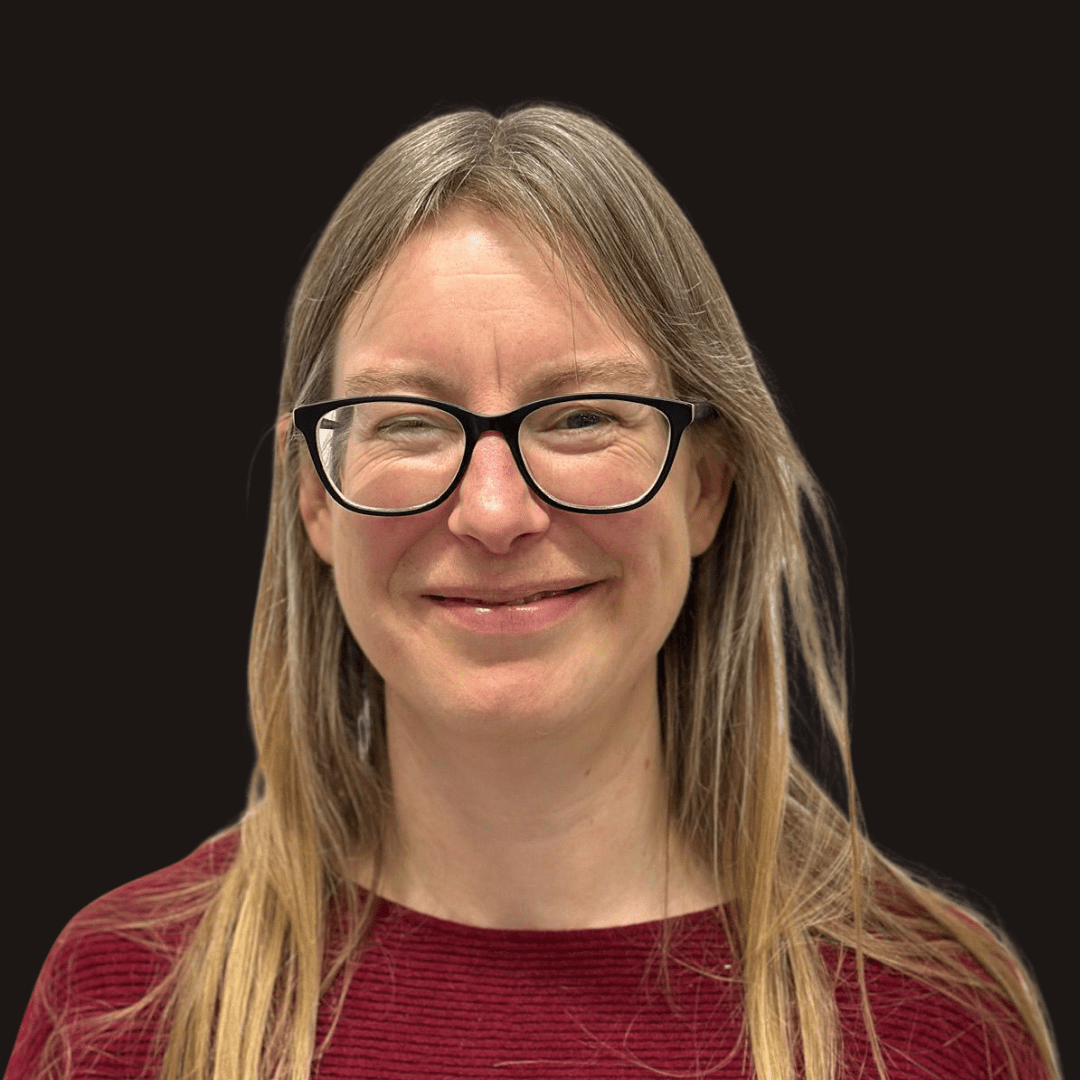 A woman with long blonde hair and glasses smiles at the camera. She is wearing a red textured top, set against a plain black background—a portrait reflecting the vibrant spirit of Ealaín na Gaeltachta.