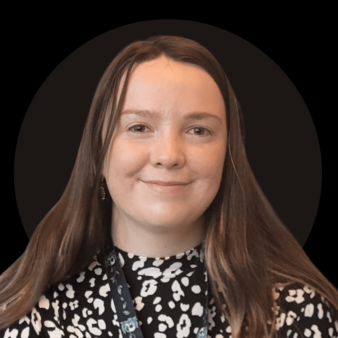 A woman with long brown hair, wearing a black and white patterned top and a dark lanyard, smiles at the camera against a plain black background—reflecting the creative spirit of Ealaín na Gaeltachta.