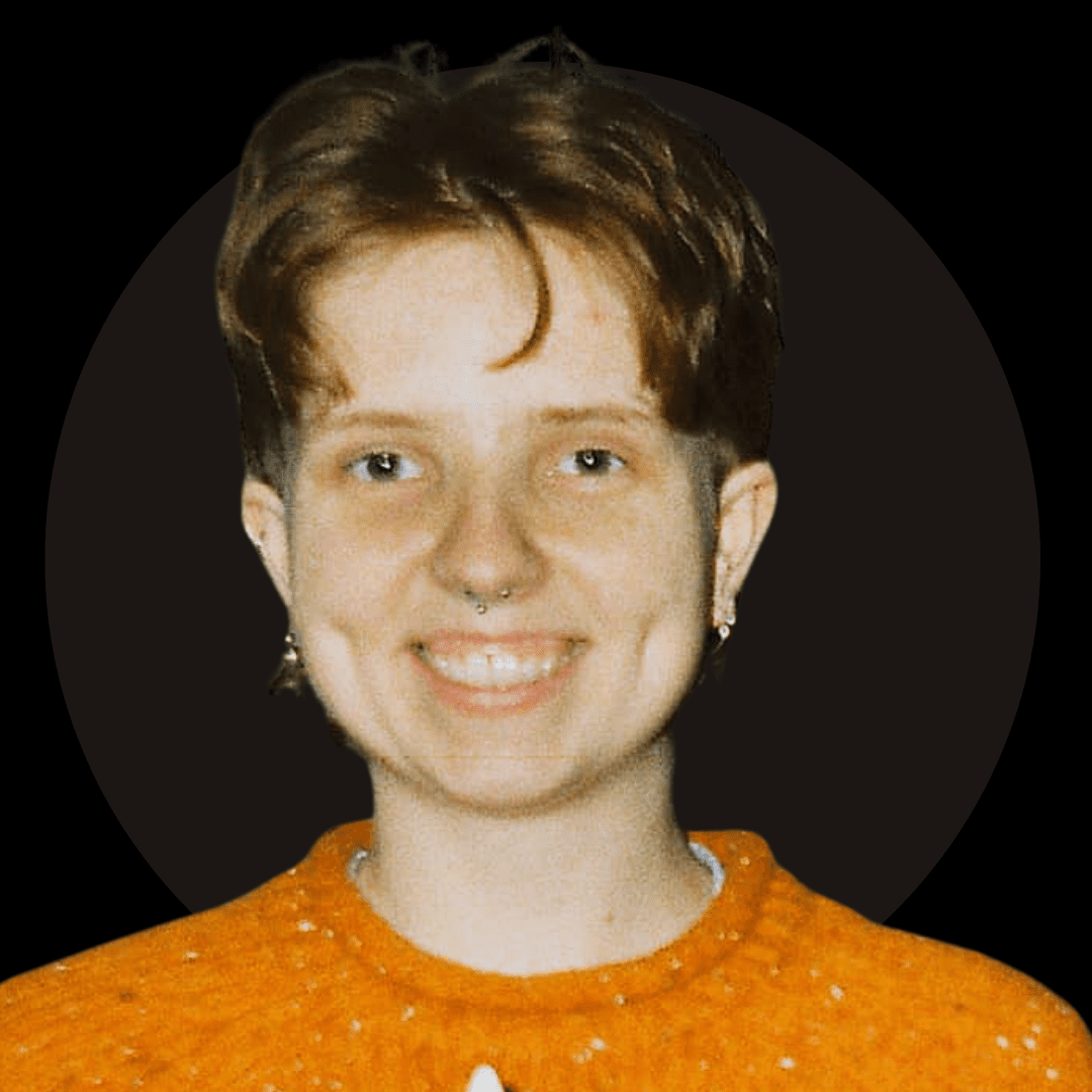 A person with short, light brown hair and a septum piercing smiles at the camera. They are wearing an orange textured jumper, small hoop earrings, and bring a modern touch to Ealaín na Gaeltachta. The black background has a faint dark circle behind their head.