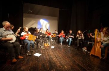 A group of musicians sits in a semicircle on a dark stage, playing various instruments including a banjo, accordion, guitar, violin, and harp—an intimate performance enriched by the spirit of Ealaín na Gaeltachta.